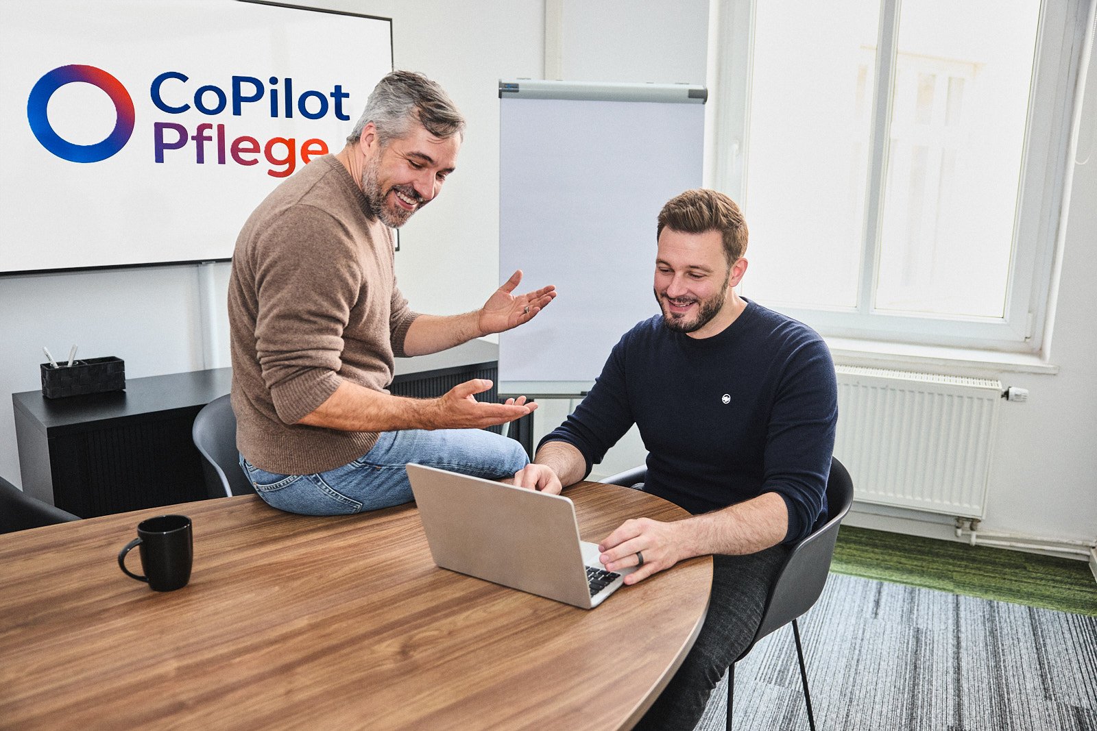 Zwei Kollegen im Gespräch am Laptop, im Hintergrund das Logo von CoPilot Pflege